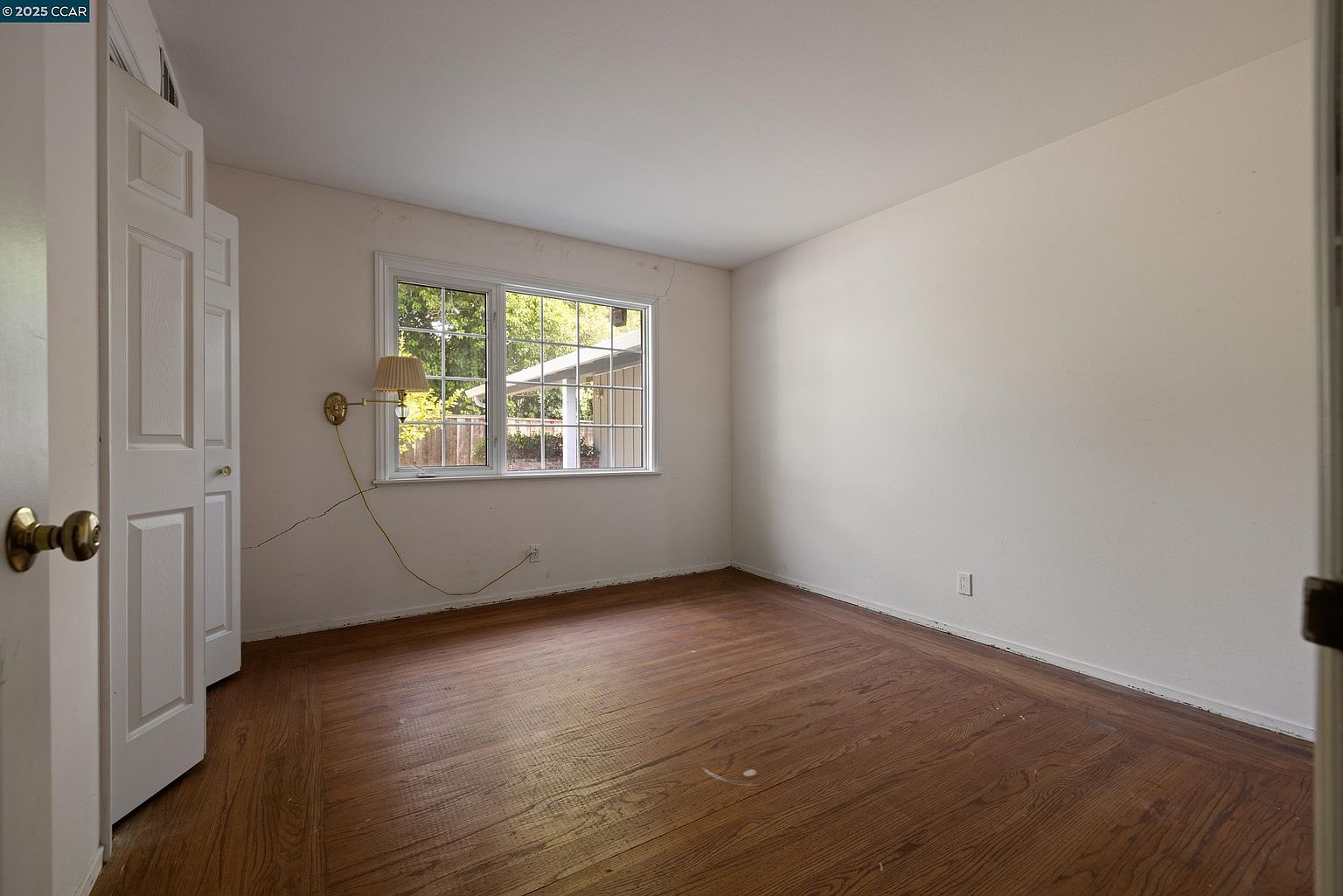This is an interior shot of a bedroom featuring hardwood floors and neutral-colored walls. A window provides natural light, and a wall-mounted lamp is visible. The room appears to be empty and ready for furnishing, offering a blank canvas for potential buyers.