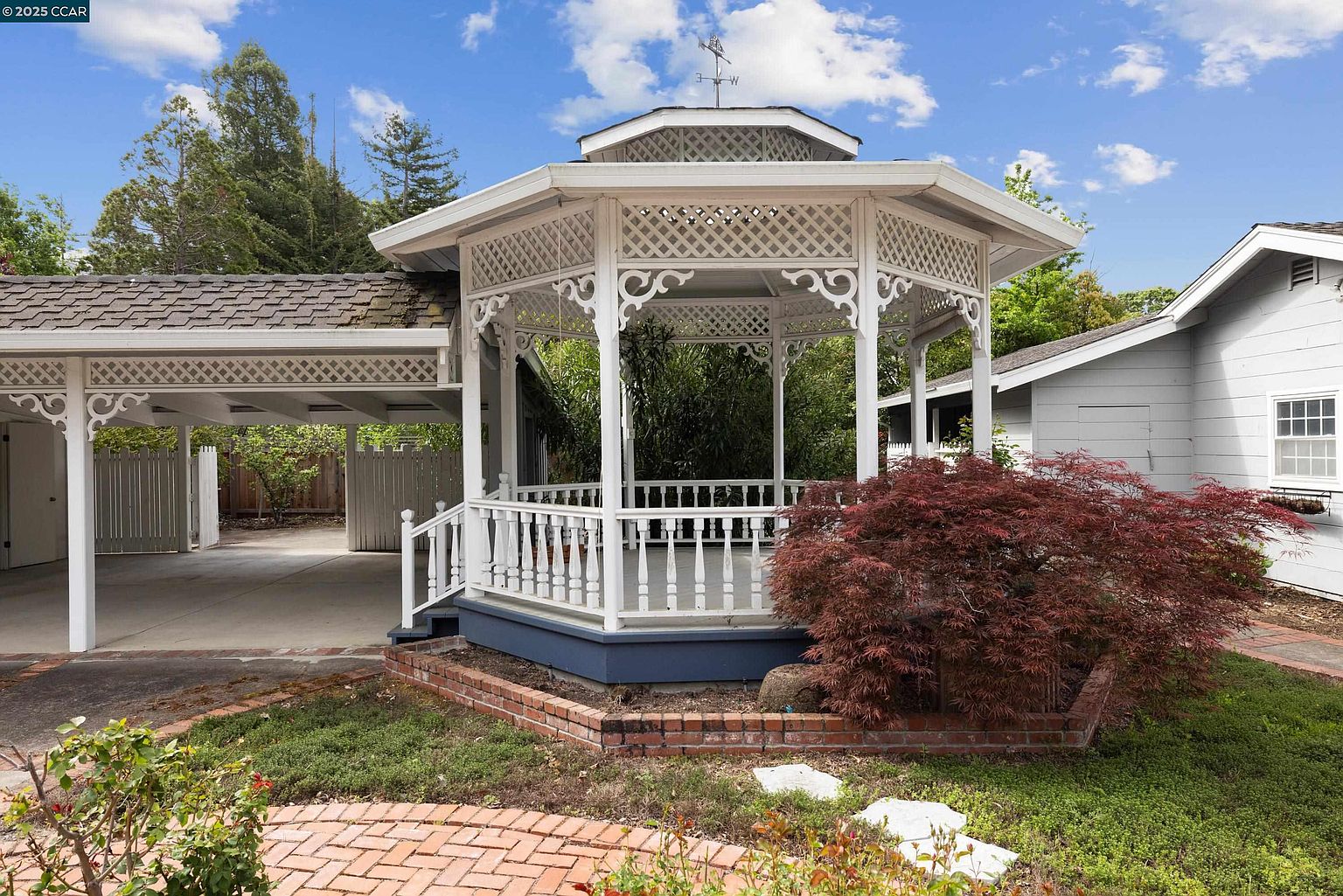 This image showcases a charming gazebo in a well-maintained yard. The gazebo features intricate latticework and a weather vane, adding a touch of elegance. A vibrant red Japanese maple tree complements the scene, while a brick pathway and lush greenery enhance the garden's appeal.