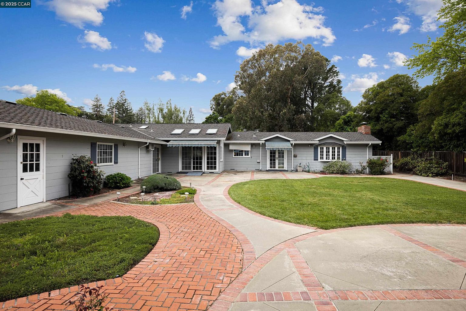 This image showcases the front exterior of a single-story home with a well-manicured lawn and a brick pathway leading to the entrance. The house features a light gray exterior, dark shutters, and a neatly maintained roof with skylights. The landscape includes mature trees and shrubs, adding to the property's curb appeal and creating a welcoming atmosphere.