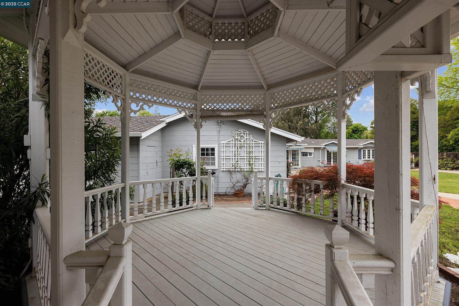 This image showcases a charming gazebo in a well-maintained yard. The gazebo features intricate latticework and a classic design, offering an inviting space for relaxation or entertainment. In the background, a quaint house with a light gray exterior and white trim adds to the picturesque setting, suggesting a peaceful and idyllic property.