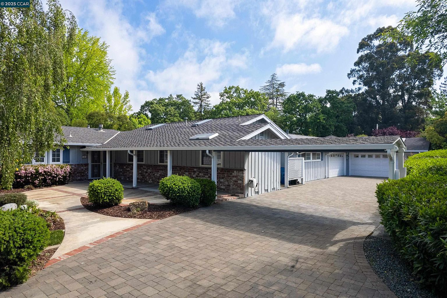 This is a front exterior view of a single-story home with a well-maintained brick driveway and mature landscaping. The house features a gray exterior with brick accents, a dark gray roof, and a covered carport leading to a garage. The overall impression is one of a charming and inviting property.