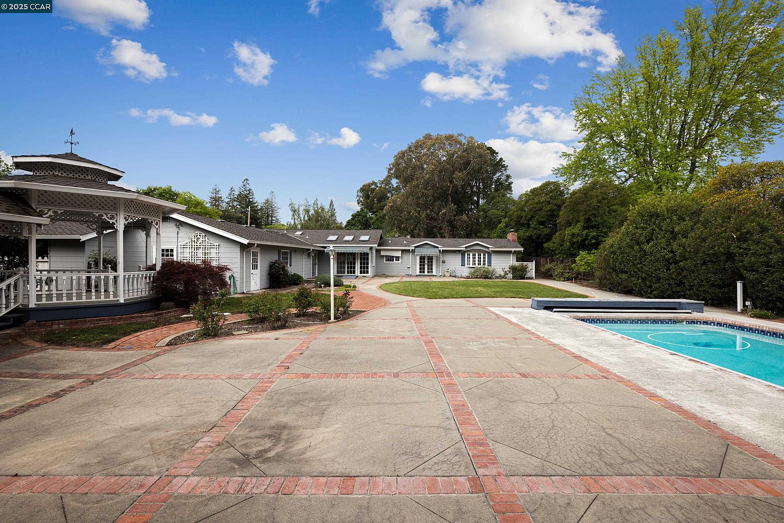 This image showcases the rear exterior of a well-maintained property, featuring a charming gazebo, a sprawling paved patio with brick accents, and a refreshing swimming pool. The house itself exhibits a classic architectural style with multiple sections, complemented by lush landscaping and mature trees that enhance the property's privacy and appeal. The perspective is from the patio, looking towards the house, creating a sense of spaciousness and inviting outdoor living.