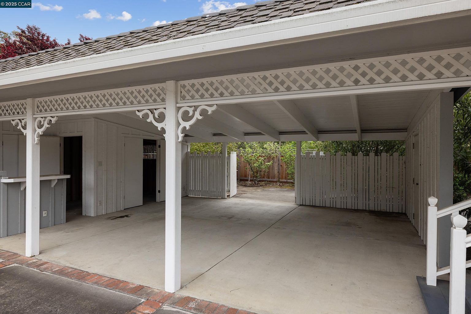 The image showcases a well-maintained carport with white support beams and decorative lattice work. The concrete floor is clean, and there are storage areas or small rooms built into the sides of the carport. The view extends to a fenced area with greenery beyond, creating a sense of openness and functionality.