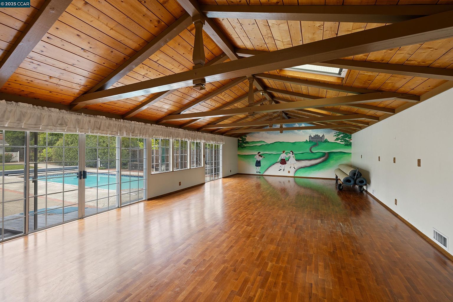 This sunroom features a wood-paneled ceiling with exposed beams and a skylight, creating a warm and inviting atmosphere. Large windows offer views of the pool area, while a mural adds a unique artistic touch to the space. The hardwood floors and ample natural light make this room a versatile and attractive feature of the property.