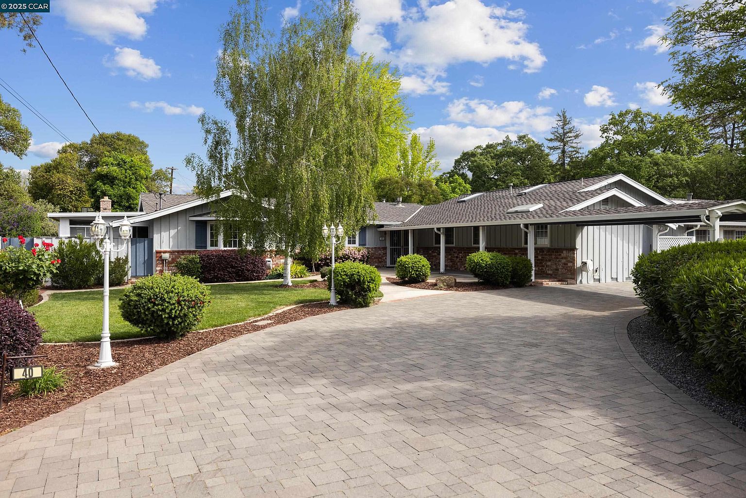 This is a front view of a single-story home with a long, paved driveway leading up to the house. The house features a combination of light gray siding and brick accents, with a gray roof and well-maintained landscaping. Mature trees and manicured bushes add to the property's curb appeal, creating a welcoming and established feel.