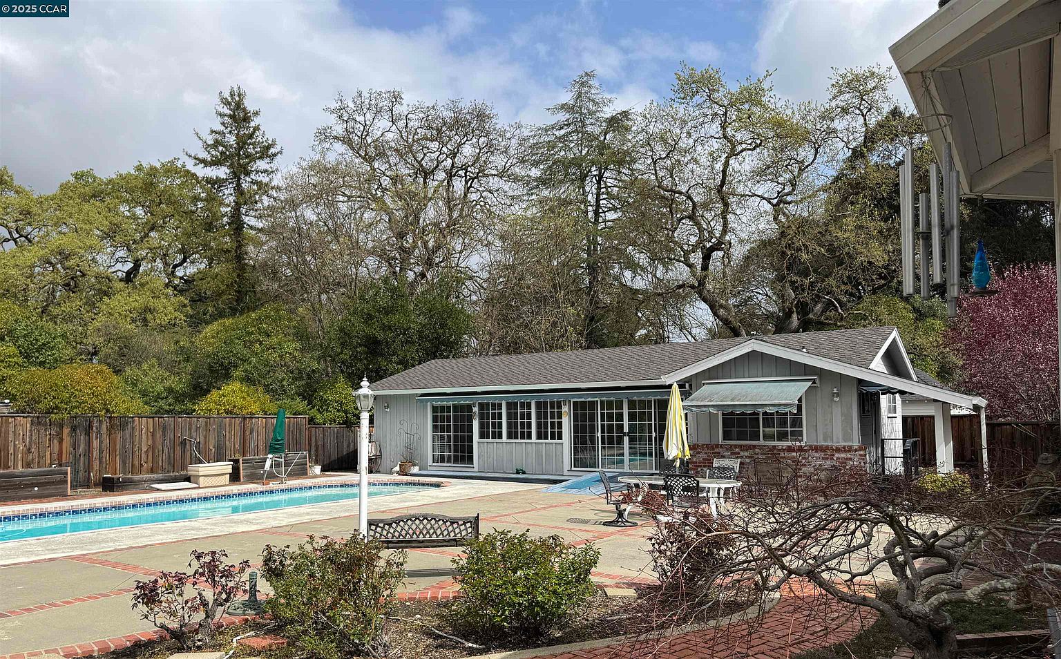 This image showcases a beautiful backyard featuring a swimming pool and patio area. The pool is surrounded by a brick patio with outdoor seating, including a bench and table with chairs. The house has a sunroom with large windows overlooking the pool area, and mature trees provide shade and privacy, creating a serene and inviting outdoor space.