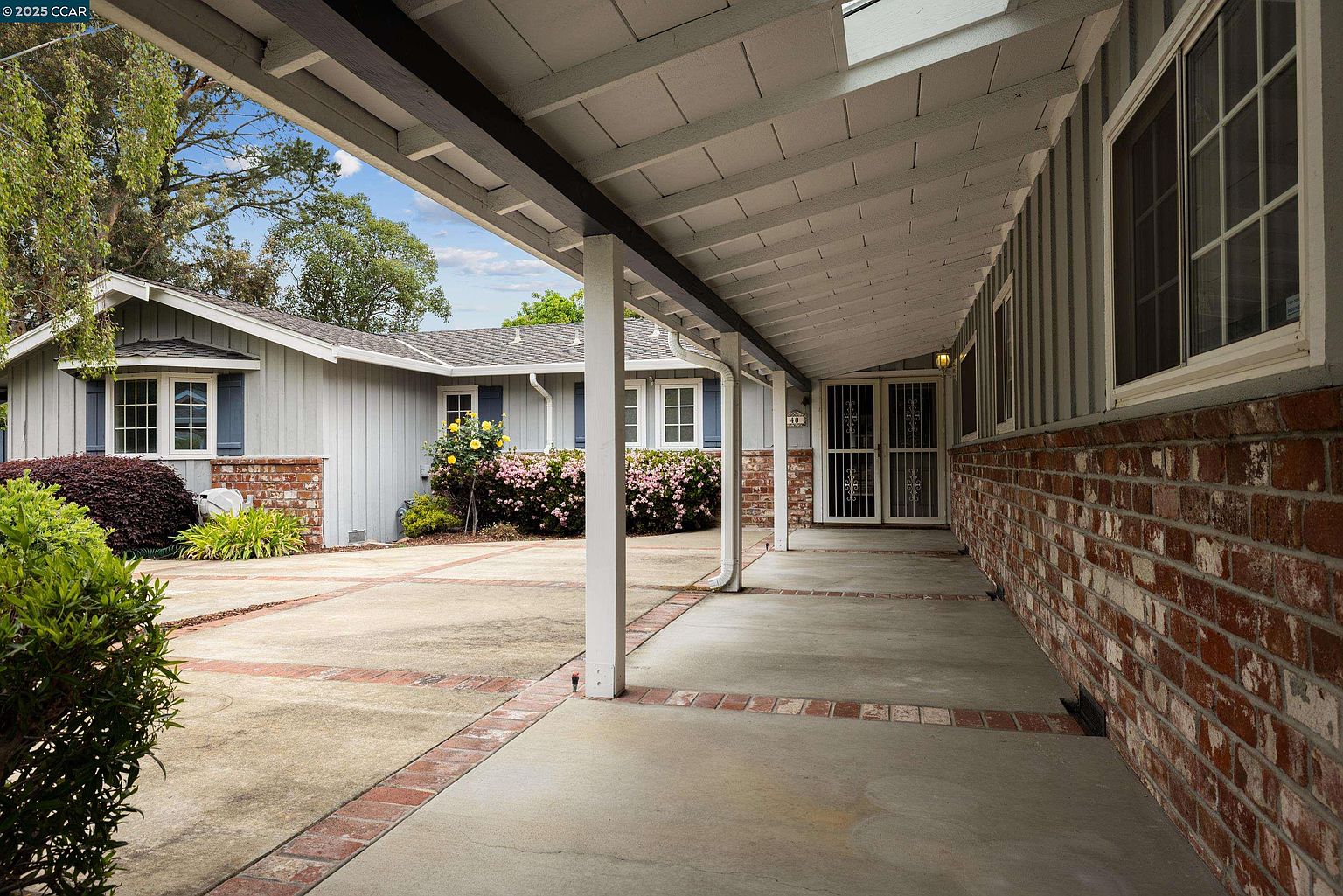 This image showcases the entryway of a charming single-story home, featuring a covered walkway with brick accents and a brick facade on the right side. The house has light gray siding and dark blue shutters, complemented by well-maintained landscaping with colorful flowers and greenery. The overall impression is inviting and well-kept.