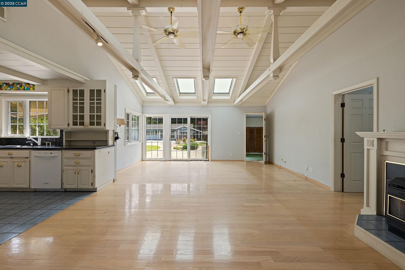 This interior shot showcases a spacious living room with a vaulted ceiling featuring exposed beams and skylights. The room transitions into a kitchen area with white cabinets and black countertops. Natural light floods the space through sliding glass doors leading to the exterior, highlighting the light hardwood flooring and creating a bright, airy atmosphere.