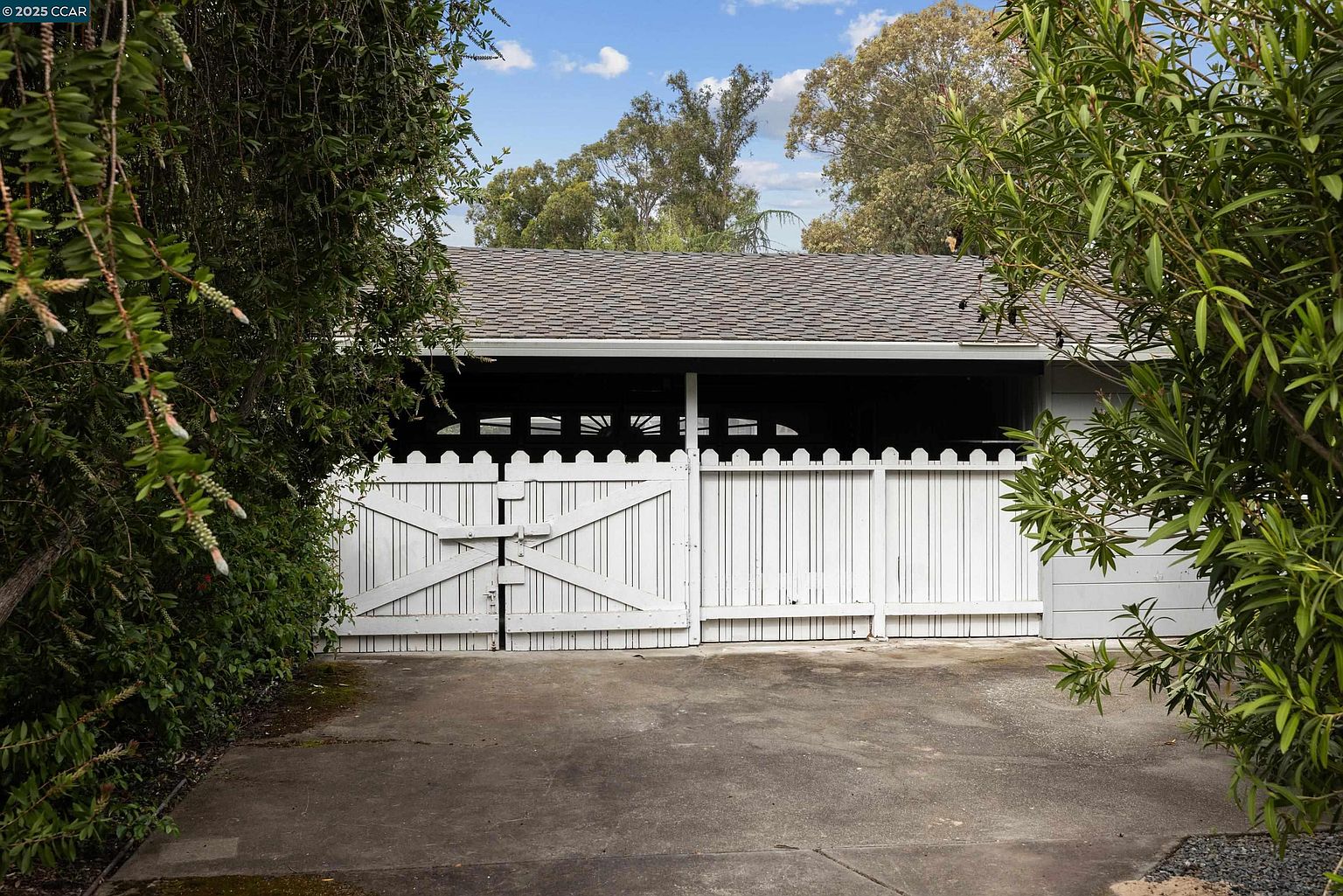 The image showcases a garage exterior with a white picket fence gate. The garage itself is painted in a neutral tone, and the roof is visible above. The driveway leads up to the garage, and greenery frames the scene on both sides, creating a sense of privacy and seclusion.