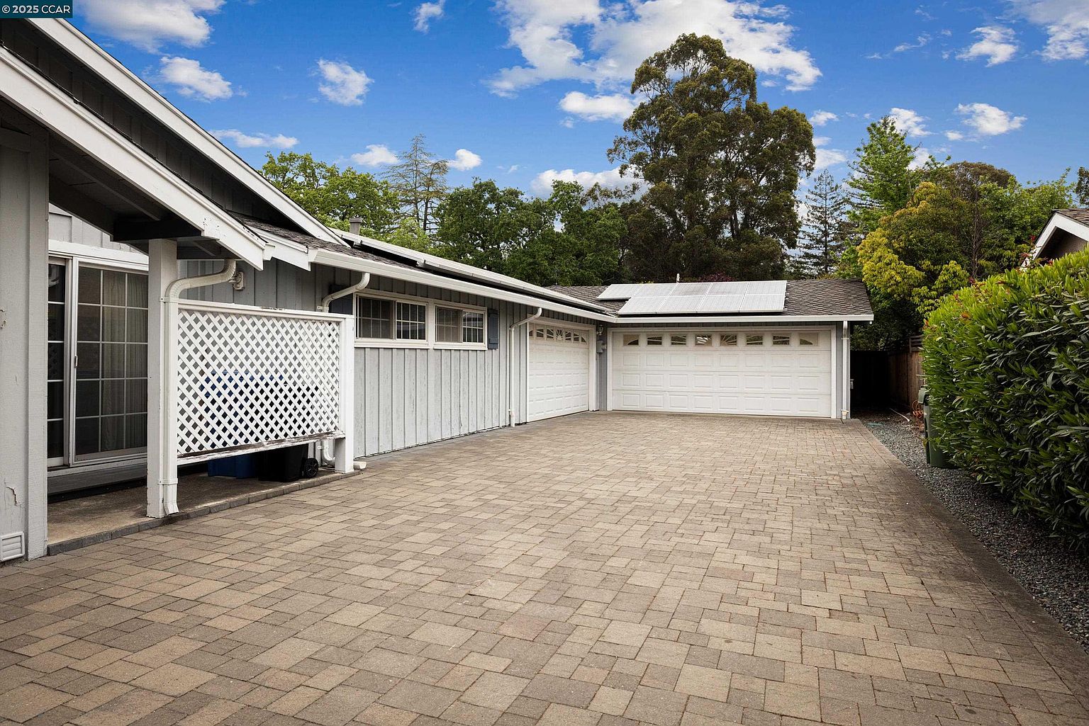 This image showcases the exterior of a home, focusing on the garage and driveway. The garage features two white doors and solar panels on the roof. The driveway is paved with interlocking bricks, leading up to the garage. The house is painted in a light gray color with white trim, and there is a white lattice fence on the left side of the house. The perspective is from the front of the house, looking towards the garage.