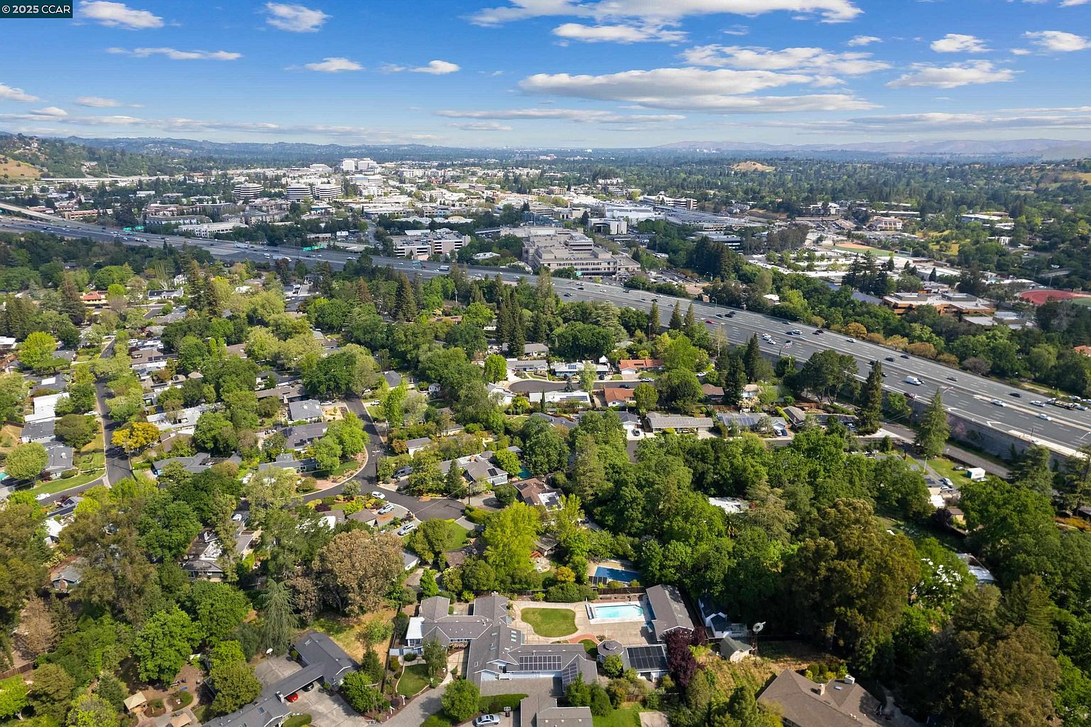This aerial view showcases a residential neighborhood with lush greenery and mature trees. A highway runs through the landscape, connecting the area to a distant cityscape. Several homes feature swimming pools, and solar panels are visible on some rooftops, highlighting energy efficiency.