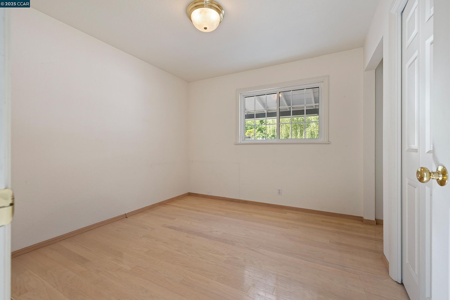 This is a bright, empty bedroom featuring light hardwood floors and white walls. A window provides natural light, and a simple ceiling light fixture illuminates the room. A white door with gold hardware is partially visible on the right, suggesting access to another area of the house.