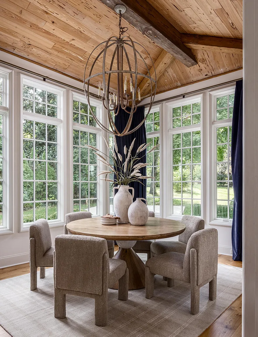 This is an interior shot of a sunlit dining room featuring a round wooden table surrounded by four upholstered chairs. A large, spherical chandelier hangs above the table, and the room is enclosed by large windows offering views of the greenery outside. The room has a warm and inviting atmosphere, enhanced by the natural light and neutral color palette.
