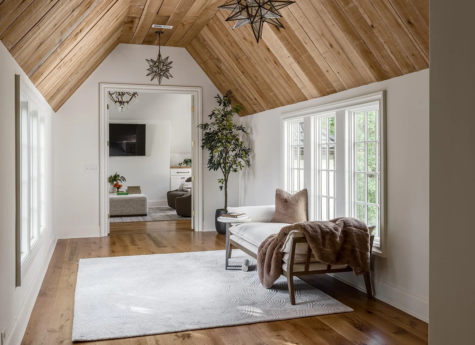 This primary bedroom features a vaulted wood plank ceiling and a neutral color palette. A large window provides ample natural light, and a chaise lounge offers a comfortable seating area. The room opens into another living space, creating an open and airy feel.