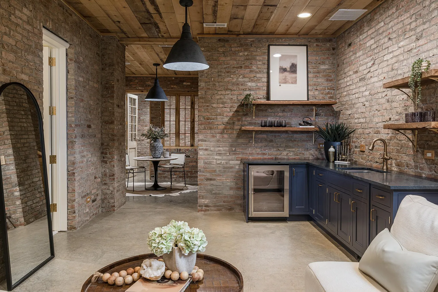 This interior shot showcases a stylish basement area with exposed brick walls and a wooden plank ceiling. A wet bar with dark blue cabinetry and a wine fridge adds a touch of luxury, while pendant lighting and wooden shelves enhance the rustic-chic aesthetic. The space also features a dining area and a glimpse of comfortable seating, creating an inviting and versatile living space.
