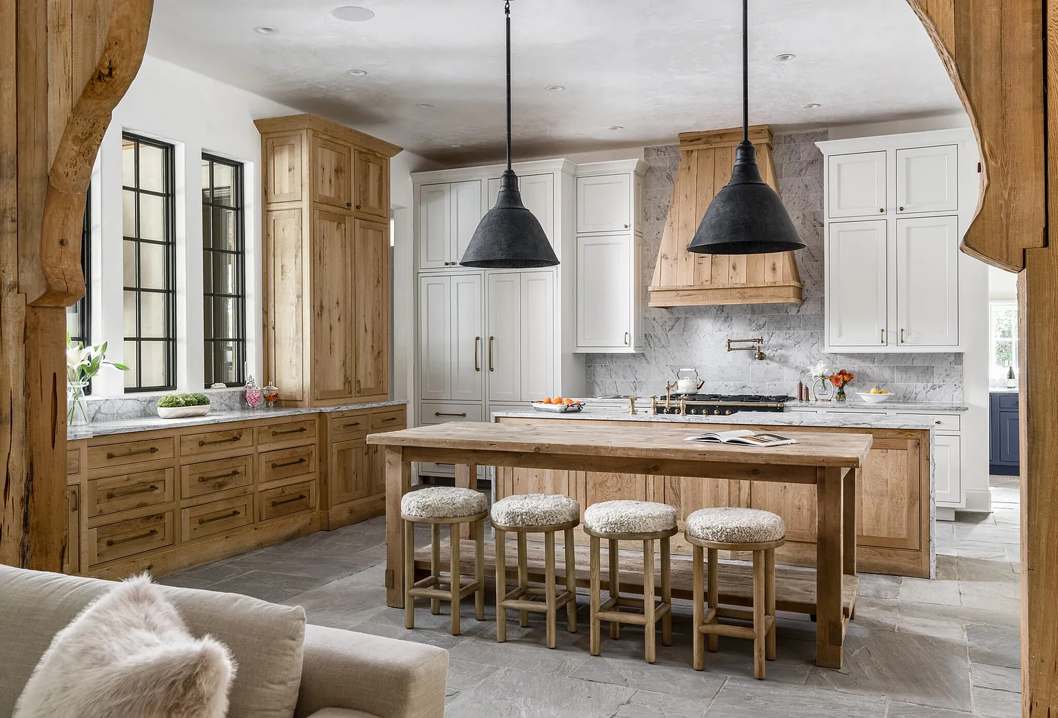 This is a well-lit, professionally staged kitchen featuring a large wooden island with seating, contrasting white and natural wood cabinetry, and modern pendant lighting. The kitchen has a rustic yet elegant feel, with stone flooring and marble countertops adding to the luxurious ambiance. The perspective is from the living room, looking into the kitchen.