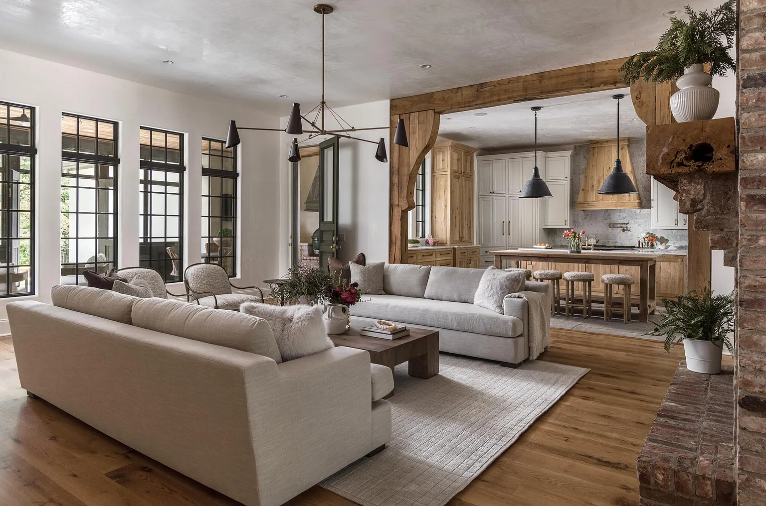 This is an interior shot of a living room that seamlessly flows into the kitchen. The living room features two large sofas, a coffee table, and large windows that provide ample natural light. The kitchen is visible through a wooden archway and includes an island with bar stools and pendant lighting, creating an open and inviting atmosphere.