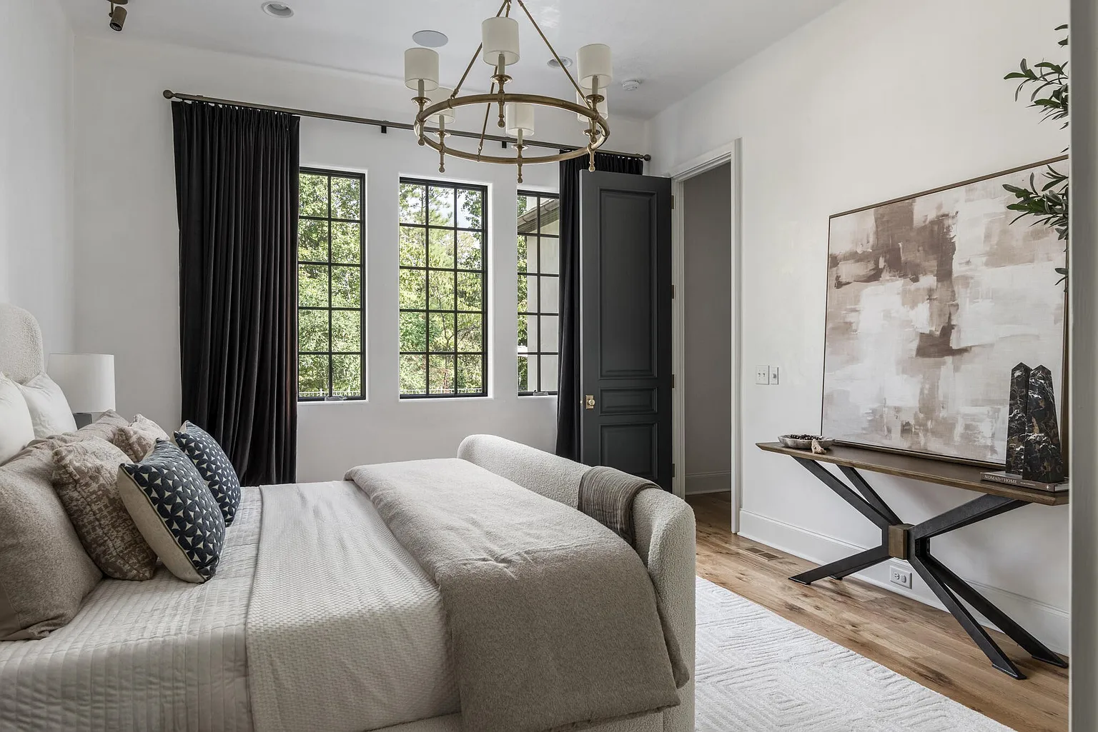 This is a well-lit primary bedroom featuring a large bed with neutral-toned bedding and decorative pillows. The room has three windows with black frames and dark curtains, providing natural light and a view of the outdoors. A modern chandelier hangs from the ceiling, and a console table with artwork adds a touch of elegance to the space.