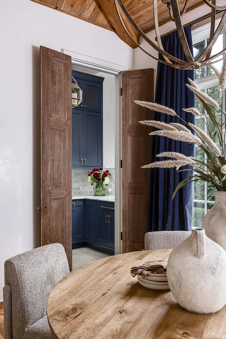 This interior shot showcases a dining room with a rustic-chic aesthetic. A round wooden table is the centerpiece, complemented by upholstered chairs and decorative vases. Through an open doorway, a glimpse of a blue-toned kitchen adds depth and interest to the scene.