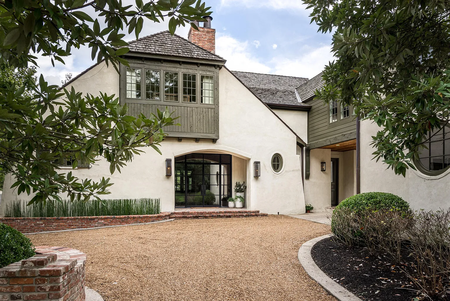 This is a front exterior view of a charming home with a gravel driveway. The house features a unique architectural style with a combination of stucco and wood siding, complemented by a brown shingle roof. A brick border adds character to the landscape, and mature trees frame the property, enhancing its curb appeal.