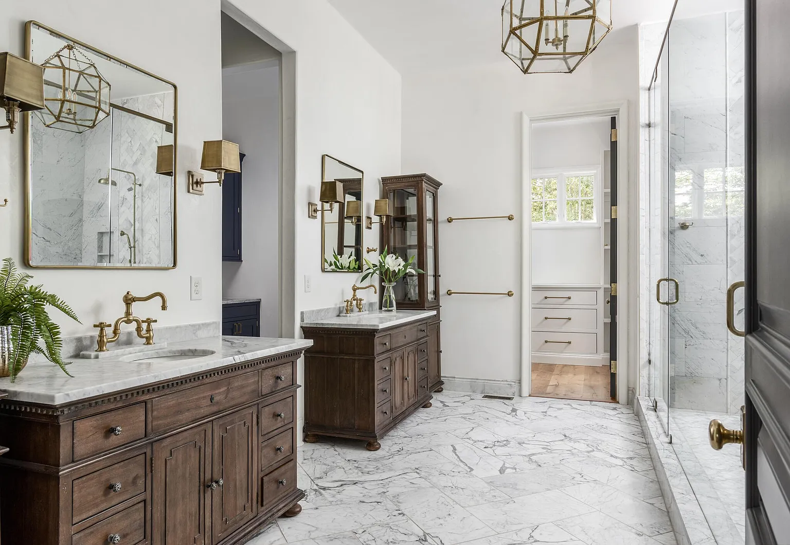 This is a luxurious primary bathroom featuring dual dark wood vanities with marble countertops and brass fixtures. The floor is covered in marble tiles, and a glass-enclosed shower is visible in the background. A walk-in closet is accessible through a doorway, and the overall style is elegant and sophisticated.