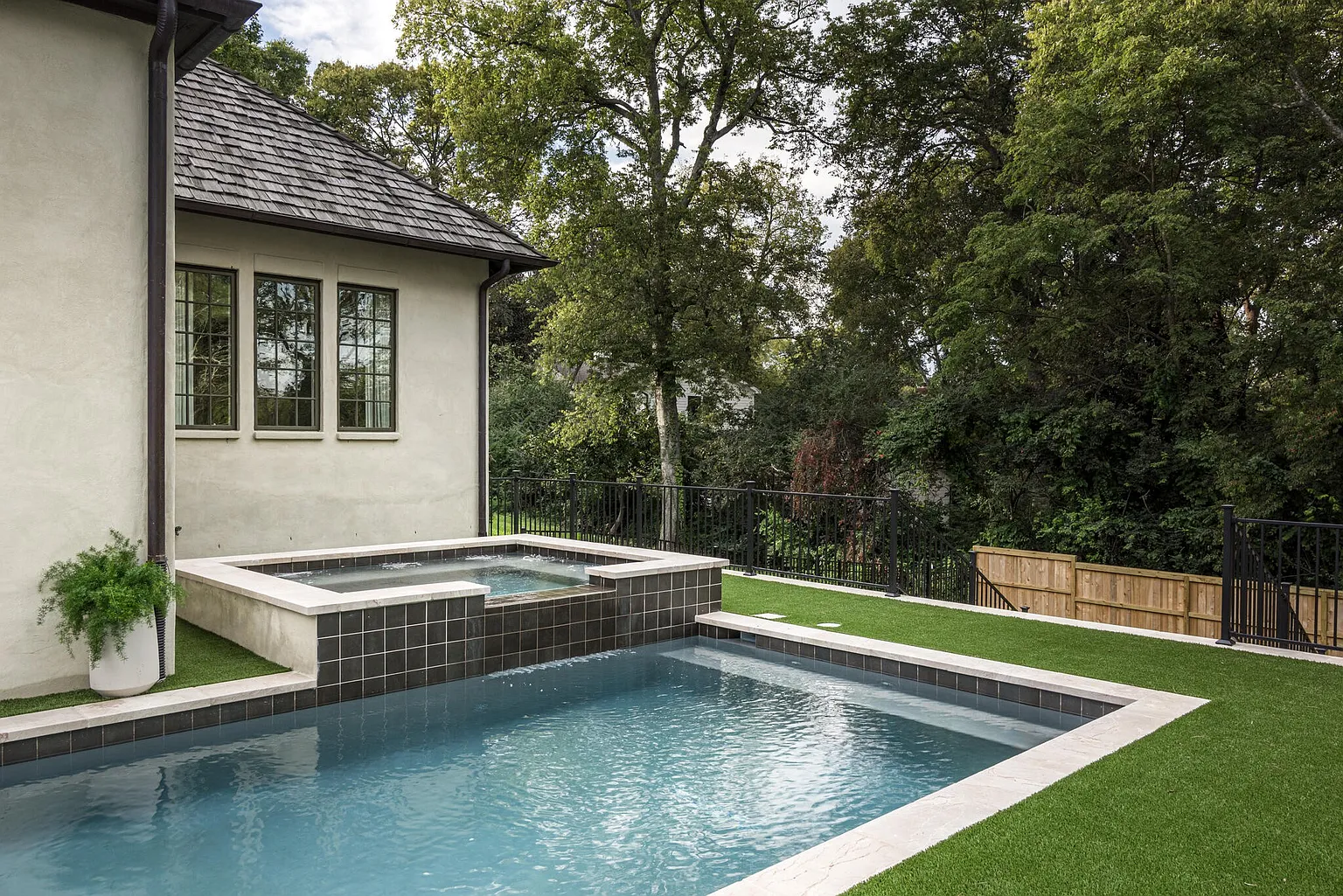 This image showcases a luxurious backyard pool and spa area. The pool features a modern design with clean lines and a light blue interior, complemented by a raised spa with dark tile accents. Artificial turf surrounds the pool, providing a low-maintenance and visually appealing space, while a black metal fence encloses the yard. The camera angle is a medium shot, capturing the pool, spa, and surrounding landscaping.