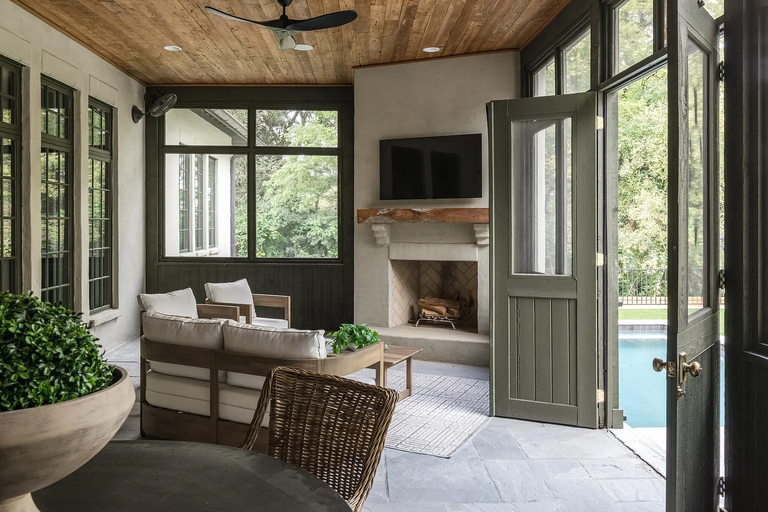 This sunroom features a cozy seating area with a fireplace and a mounted television. The room has large windows and doors that open to the outside, providing ample natural light and a view of the pool. The ceiling is wood paneled, and the flooring is stone, creating a warm and inviting atmosphere.