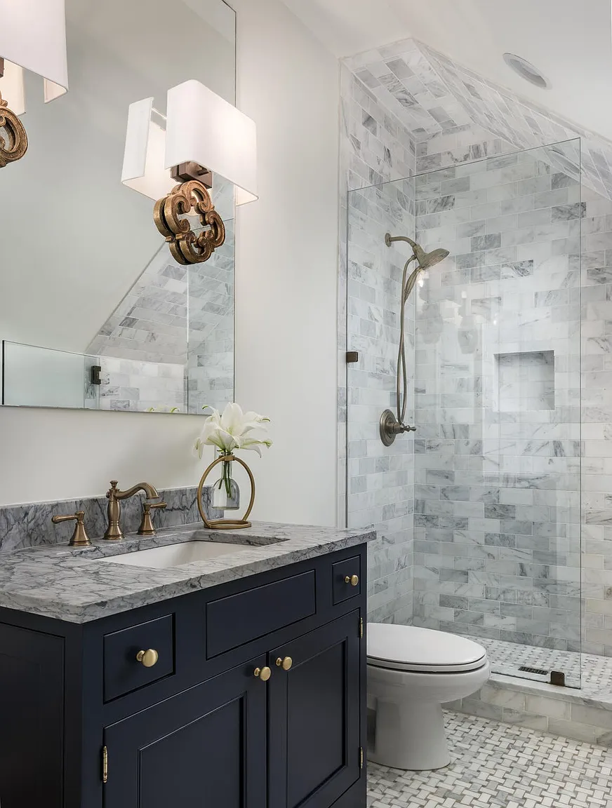 This is a well-lit bathroom featuring a dark blue vanity with a marble countertop and gold hardware. A shower with glass doors and marble tiling is visible in the background, along with a toilet. The overall impression is clean, modern, and luxurious.