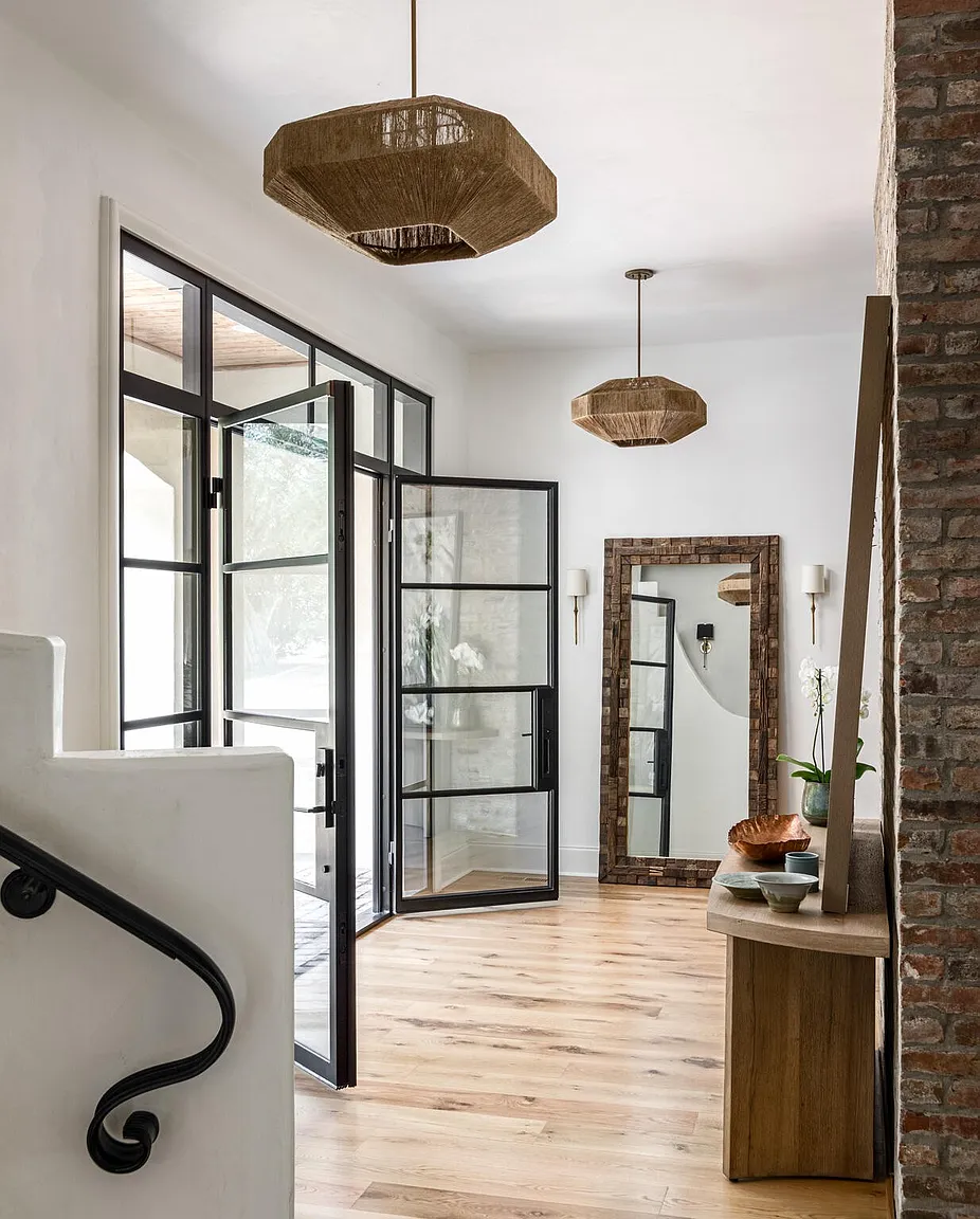 This interior shot showcases a stylish hallway with a blend of modern and rustic elements. A black-framed glass door allows natural light to flood the space, complemented by unique pendant lighting. The hallway features hardwood floors, a brick accent wall, and a staircase with a wrought iron railing, creating an inviting and sophisticated entryway.