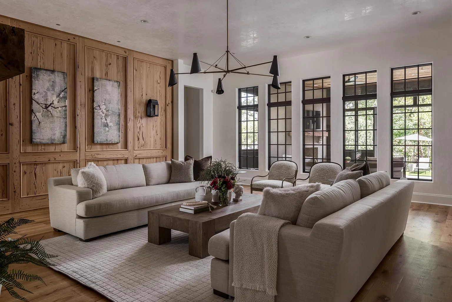 This is a beautifully designed living room featuring two large sofas, a rustic wooden coffee table, and a striking chandelier. The room is characterized by a blend of natural wood paneling and white walls, complemented by large windows that provide ample natural light. The overall aesthetic is sophisticated and inviting, creating a warm and comfortable atmosphere.