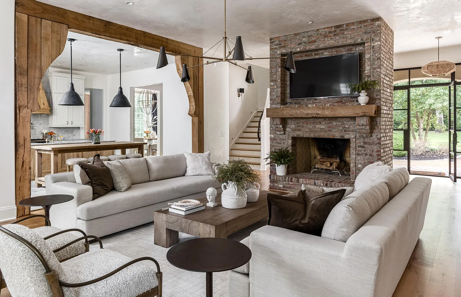 This is a warm and inviting living room featuring two light-colored sofas, a brick fireplace with a mounted television, and a rustic wooden coffee table. The room is well-lit with natural light and a modern chandelier, and the open floor plan connects to the kitchen through a wooden-framed opening. The overall style is a blend of rustic and contemporary, creating a comfortable and stylish space.