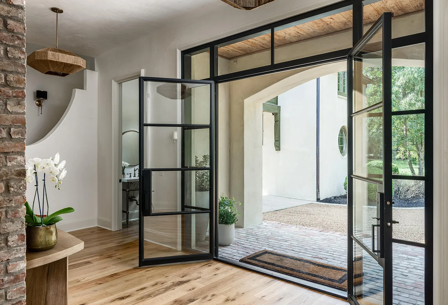 This image showcases a stylish entryway with a focus on natural light and modern design. The open black-framed glass doors lead to an exterior courtyard, creating a seamless transition between indoor and outdoor spaces. Inside, hardwood floors, a brick accent wall, and a unique light fixture add character and warmth to the space.