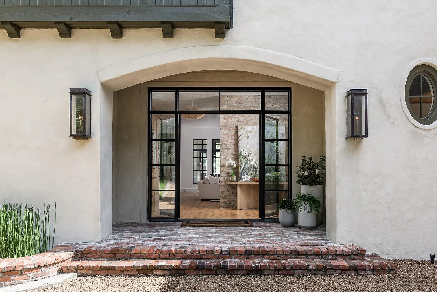This is a welcoming entryway featuring a brick staircase leading to a large arched doorway with black framed glass doors. The exterior walls are textured stucco, complemented by elegant lantern-style sconces on either side of the entrance. The overall impression is one of sophisticated charm and inviting warmth.