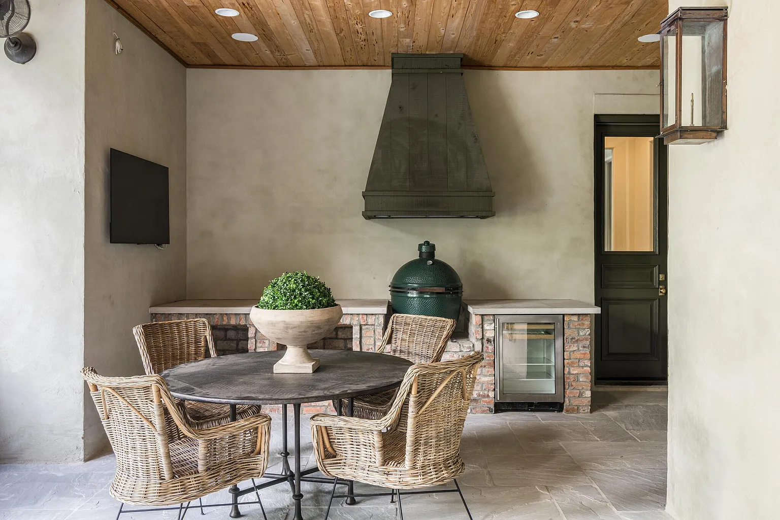 This image showcases a well-appointed outdoor patio area, featuring a round dining table surrounded by wicker chairs. The space includes an outdoor kitchen setup with a built-in grill, a beverage refrigerator, and a custom vent hood. The patio is covered with a wooden ceiling and stone flooring, creating a cozy and functional outdoor living space.