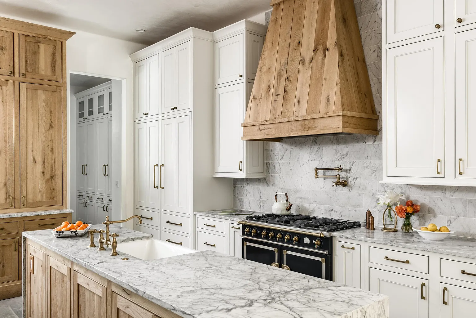 This is a well-lit kitchen featuring a large island with a marble countertop and a farmhouse sink. The cabinets are a mix of white and natural wood, complemented by brass hardware. A black range with a custom wooden hood adds a focal point, and marble backsplash completes the luxurious look.