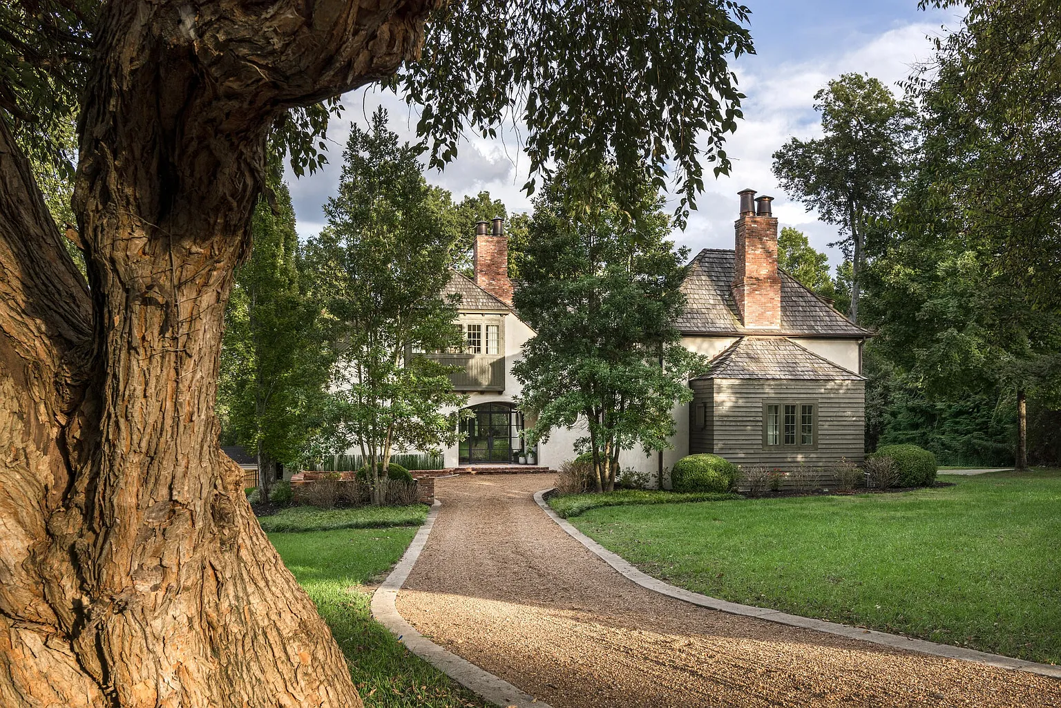 This image showcases the front exterior of a charming home, framed by a large, mature tree in the foreground. A gravel driveway leads to the house, which features a mix of white stucco and wood siding, complemented by a shingle roof and brick chimneys. The landscaping is lush and well-maintained, creating a welcoming and picturesque setting.