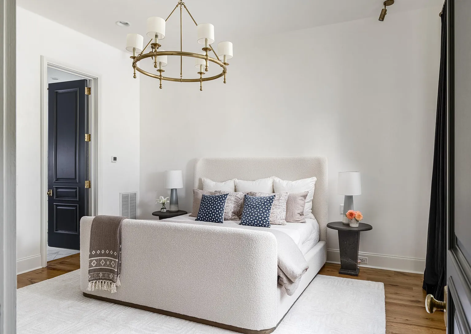 This is a beautifully styled primary bedroom featuring a plush, upholstered bed with decorative pillows and a cozy throw. The room is illuminated by a modern chandelier and bedside lamps, creating a warm and inviting atmosphere. The hardwood floors and neutral color palette contribute to a sophisticated and relaxing ambiance.
