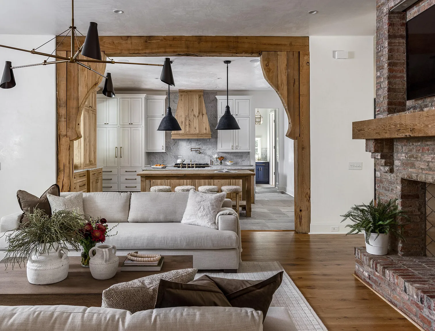 This is an interior shot of a living room that seamlessly transitions into a kitchen area. The living room features comfortable sofas, a wooden coffee table, and a brick fireplace, creating a cozy and inviting atmosphere. The kitchen, visible through a wooden archway, showcases white cabinetry, a wooden island, and modern pendant lighting, blending rustic charm with contemporary design.