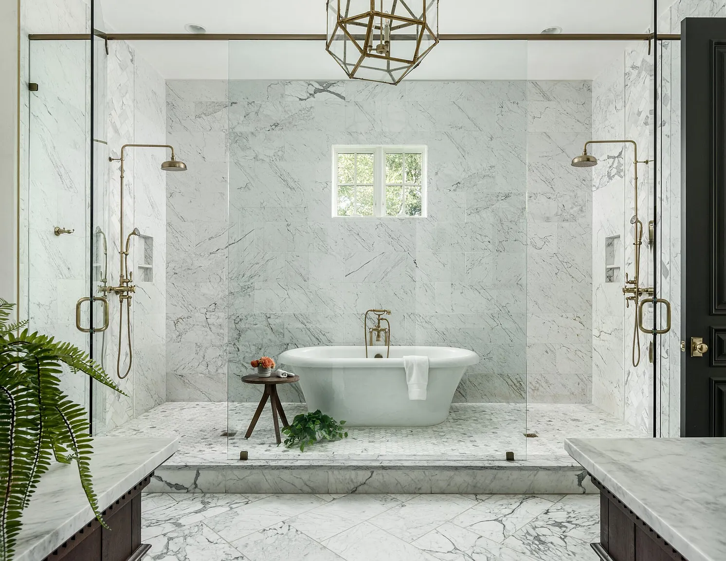 This is a luxurious primary bathroom featuring marble walls and flooring, a freestanding soaking tub, and dual shower heads enclosed by glass panels. The space is illuminated by a modern geometric chandelier, and a small window provides natural light. The overall impression is one of elegance and spa-like tranquility.