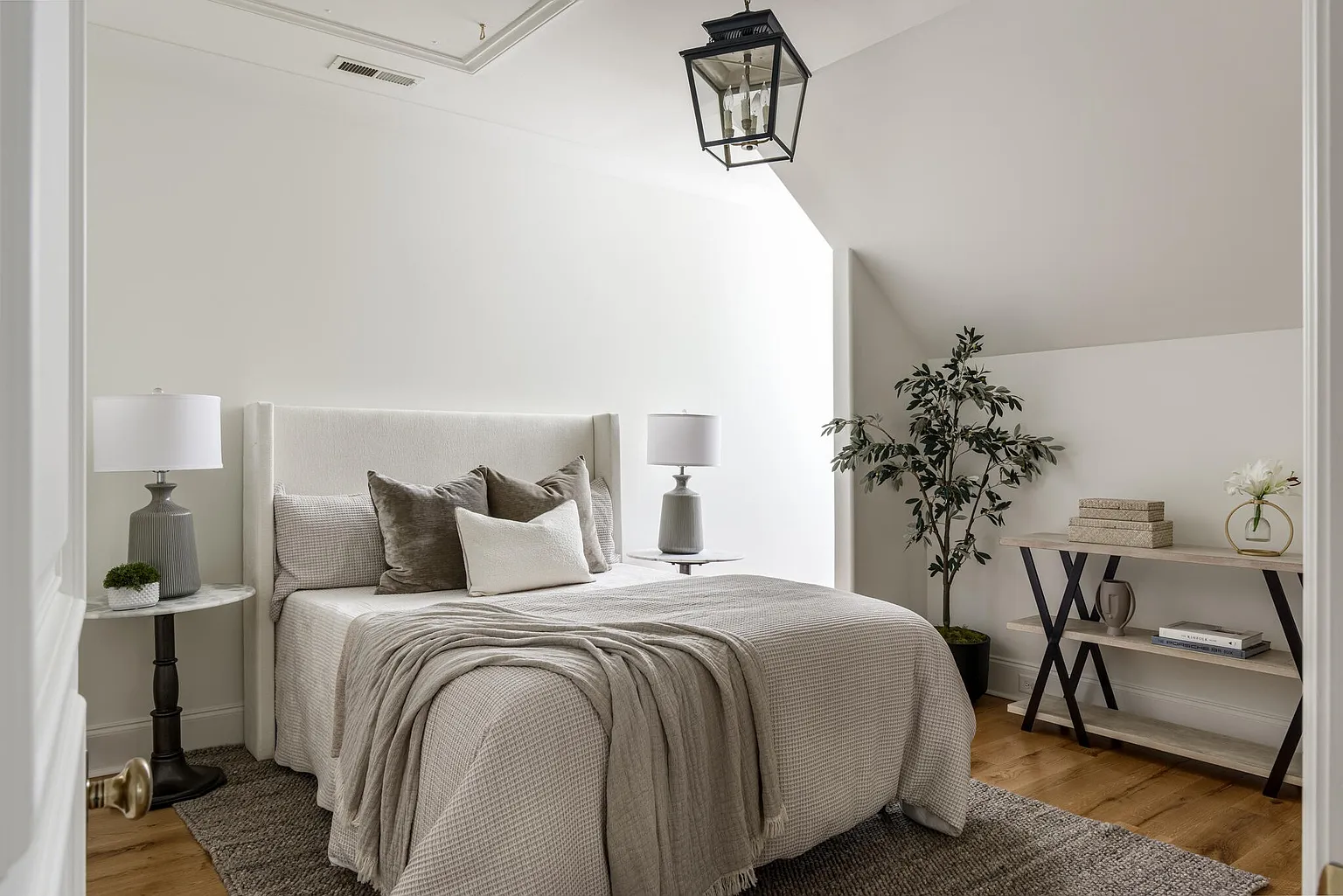 This is a well-lit bedroom featuring a neutral color palette. The bed is neatly made with a textured blanket and several pillows, and there are side tables with lamps on either side. A decorative tree and a console table with decorative items add to the room's appeal, creating a cozy and inviting atmosphere.
