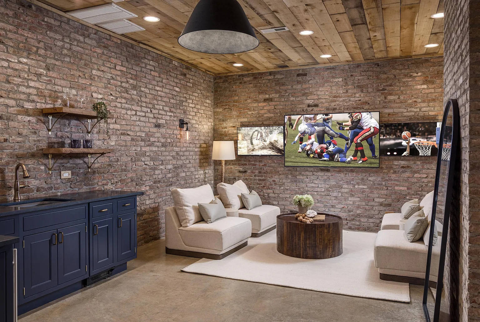 This interior shot showcases a stylish living room with exposed brick walls and a rustic wooden ceiling. The room features comfortable seating arrangements, including armchairs and a sofa, centered around a wooden coffee table. A wet bar with dark blue cabinetry adds a touch of luxury, while multiple TVs provide entertainment, creating a cozy and inviting atmosphere.