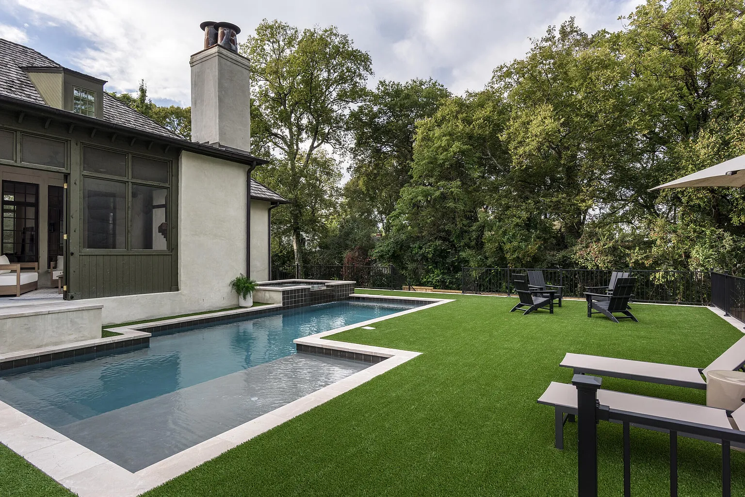 This image showcases a luxurious backyard pool and spa area. The pool features a modern design with clean lines and a dark tile finish, surrounded by a light-colored stone border. Artificial turf creates a lush green space, furnished with lounge chairs and Adirondack chairs, perfect for relaxation and entertainment. The property is surrounded by mature trees, providing privacy and a serene atmosphere.