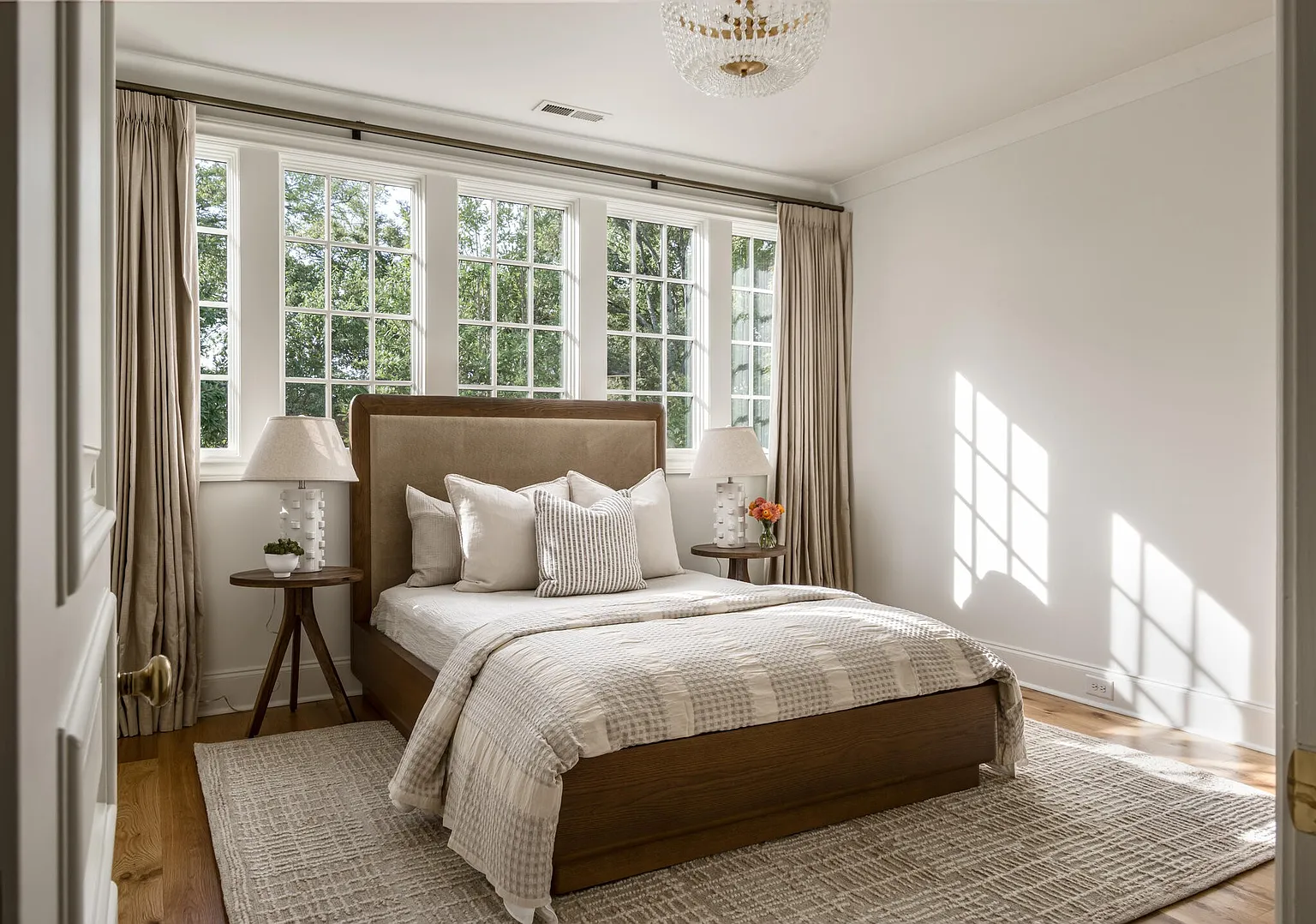 This is a bright and inviting primary bedroom featuring a large window with natural light. The room is furnished with a wooden bed frame, a neutral-toned headboard, and matching side tables with lamps. A patterned rug anchors the space, creating a cozy and serene atmosphere.