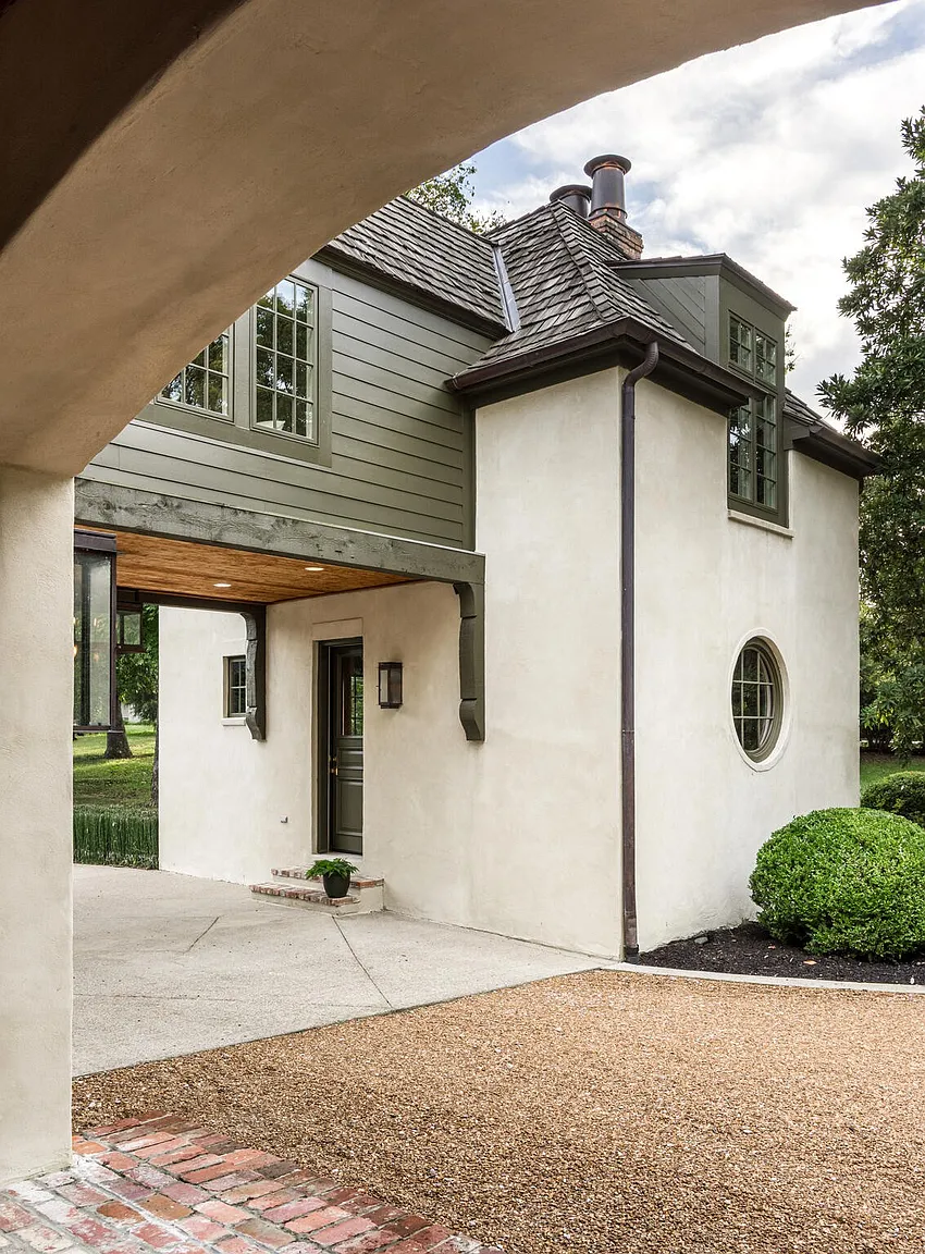 This image showcases the front exterior of a charming home, featuring a blend of textures with stucco walls, wood siding, and a shingled roof. An arched entryway leads to a covered area, and a round window adds a unique architectural detail. The landscaping includes gravel and a neatly trimmed bush, contributing to the property's curb appeal.
