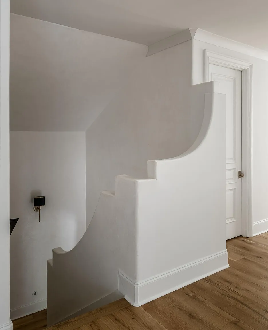 This interior shot showcases a unique staircase design with curved walls and integrated steps, all painted in a crisp white. The hardwood flooring adds warmth to the space, while a nearby door with classic paneling suggests a traditional touch. A wall-mounted sconce provides subtle lighting, enhancing the architectural details.