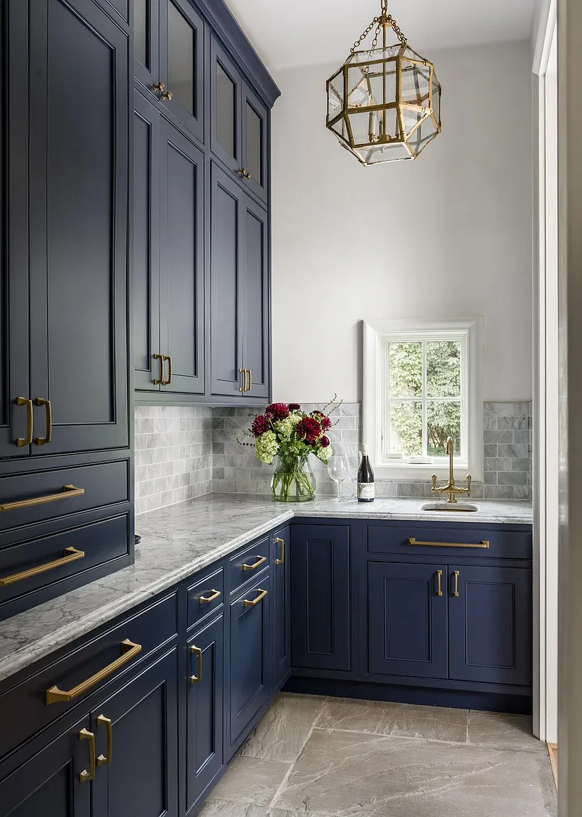 This is an interior shot of a laundry room featuring navy blue cabinetry with gold hardware, marble countertops, and a geometric gold light fixture. The room has a clean and sophisticated design, with a window providing natural light and a view of the outdoors. The overall impression is one of luxury and functionality.