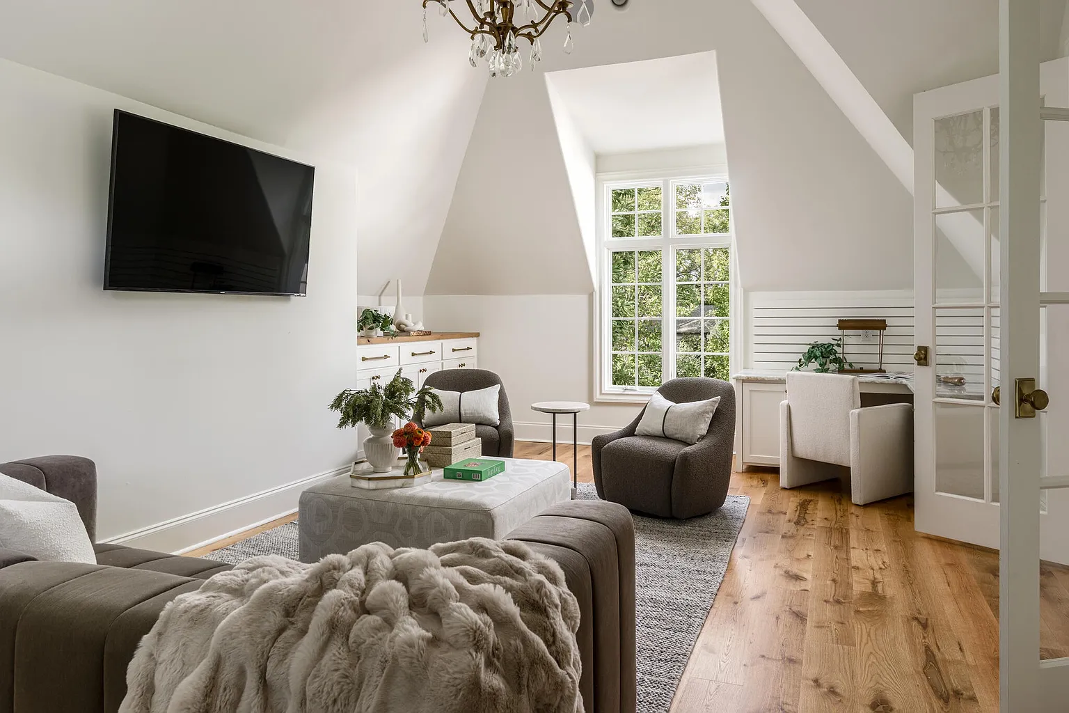 This is an inviting living room featuring a comfortable seating area with a plush sofa and two armchairs, centered around a patterned ottoman. The room is well-lit by a large window and a chandelier, and a built-in desk area is visible in the background. The hardwood floors and neutral color palette create a warm and sophisticated atmosphere.
