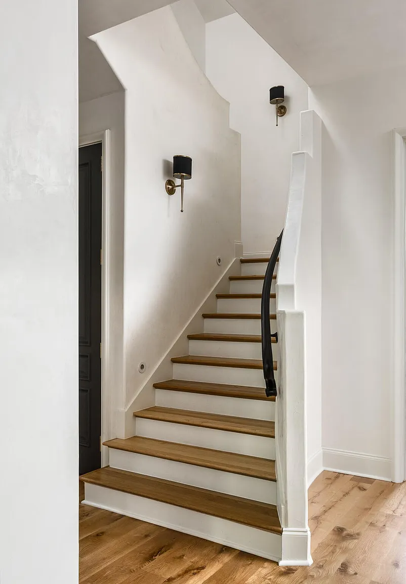 This interior shot showcases a staircase with wooden treads and white risers, complemented by a black handrail and white walls. Two sconce lights illuminate the wall, adding a touch of elegance to the space. The hardwood flooring on the lower level adds warmth and continuity to the design.