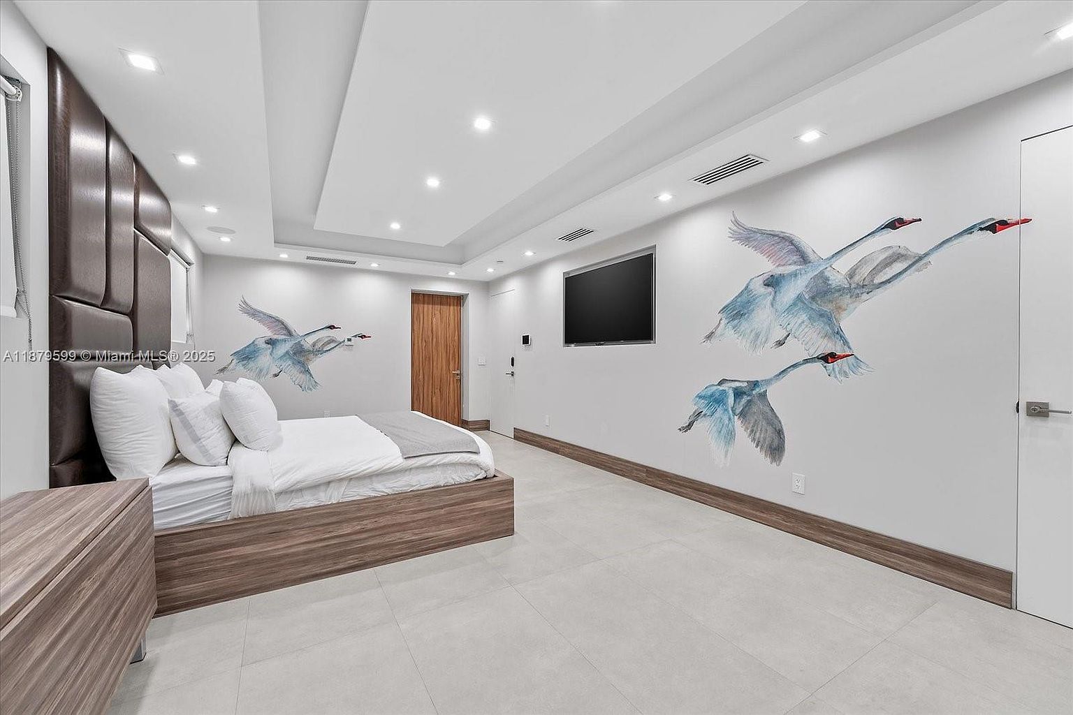 This is a modern bedroom featuring a platform bed with a padded headboard and white linens. The walls are adorned with swan artwork, and a flat-screen TV is mounted on the wall. The room has recessed lighting and a clean, minimalist aesthetic, creating a serene and stylish space.