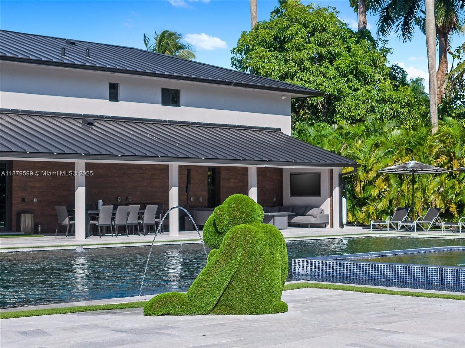 This image showcases a luxurious backyard pool area. A modern house with a dark metal roof and a covered patio with outdoor seating overlooks the pool. A unique green topiary sculpture sits beside the pool, adding an artistic touch to the landscape. The pool features a tiled edge and is surrounded by a paved patio and lush greenery, creating a serene and private oasis.